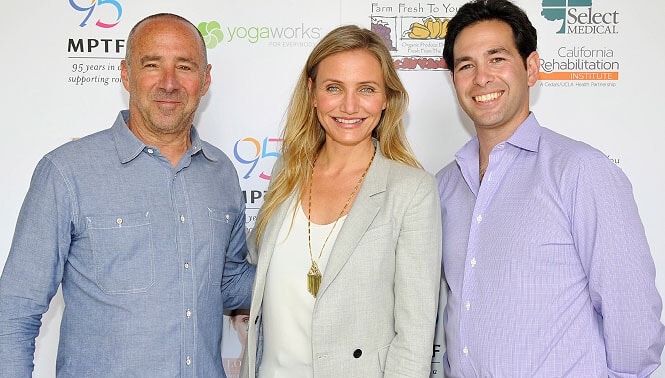 Three individuals are posing for a photo in front of a backdrop with various logos. The two men are on either side of Cameron Diaz, who is standing in the middle.