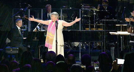 A singer stands on stage with arms outstretched, wearing a white dress and colorful scarf, while a pianist and other musicians play in the background.