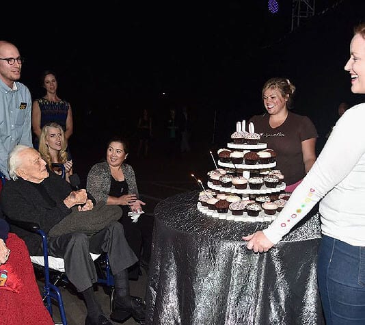 A group of people, including an older man in a wheelchair, gather around a multi-tiered stand of cupcakes with lit candles during a celebration. One woman presents the cupcakes.