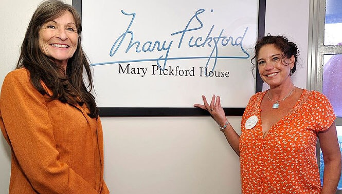 Two women standing next to a framed sign that says "Mary Pickford, Mary Pickford House." One is wearing an orange coat, and the other is in an orange patterned blouse.