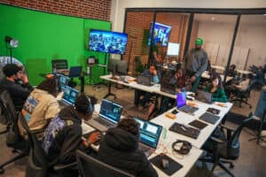 A group of people sitting at desks with laptops in front of a green screen.