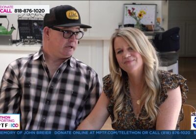 A man wearing a black and yellow cap sits next to a woman with long blonde hair; both appear on a telethon broadcast with informational text and donation numbers displayed on the screen.