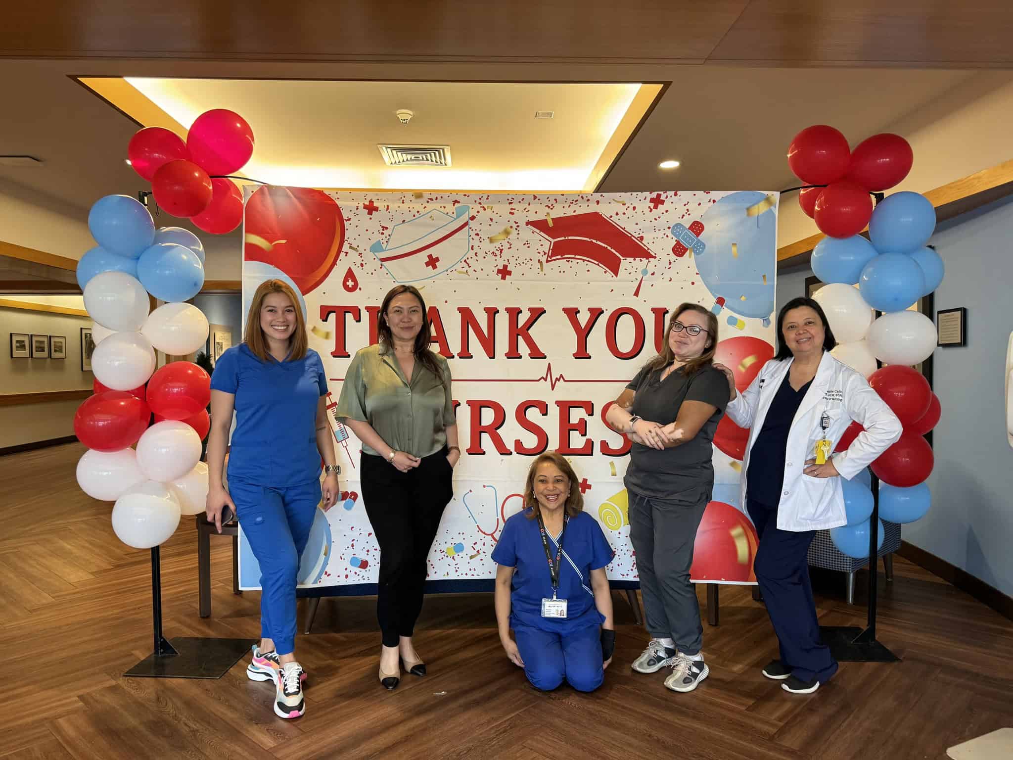 Five healthcare professionals are standing in front of a "Thank You Nurses" banner decorated with balloons. Four are standing and one is kneeling, all smiling.