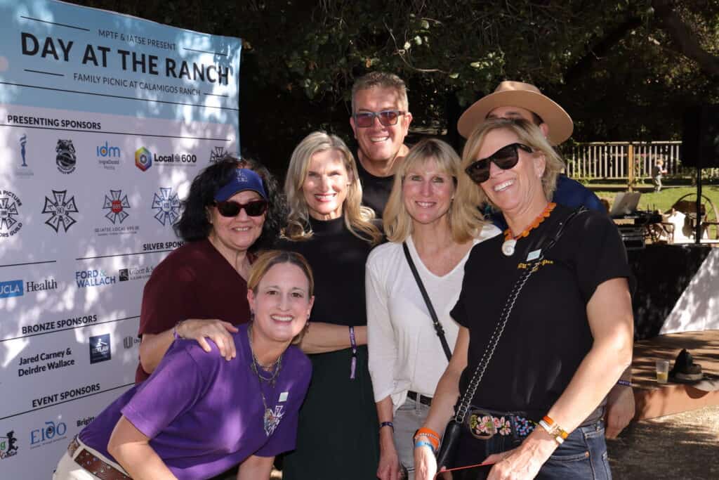 A group of people smiling at a daytime outdoor event, standing in front of a sign with sponsors' logos.