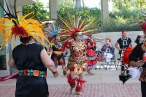 A group of people in vibrant, feathered costumes perform a traditional dance outdoors, while onlookers watch from the background.